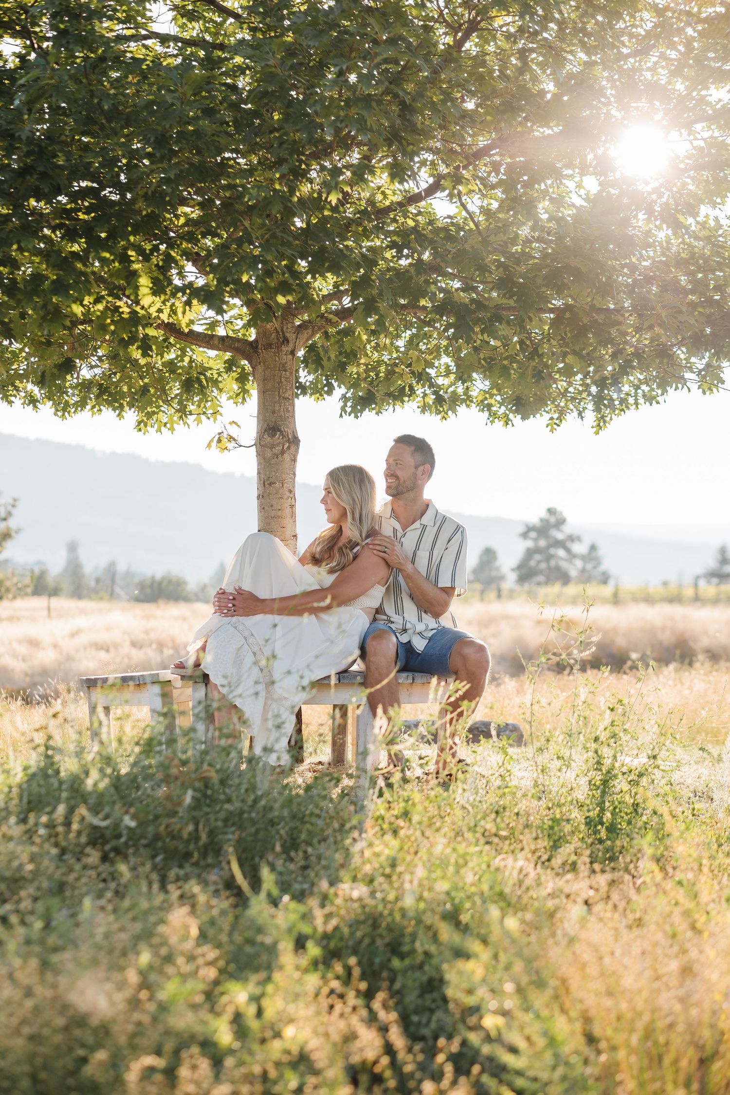 A couple sits together on a wooden swing in a sunlit meadow during golden hour with trees in the background.