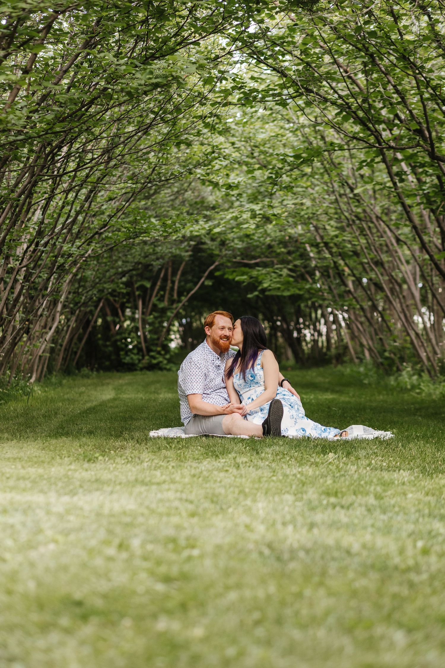 Couple lounging together on grass under a canopy of lush green trees in a romantic outdoor setting.