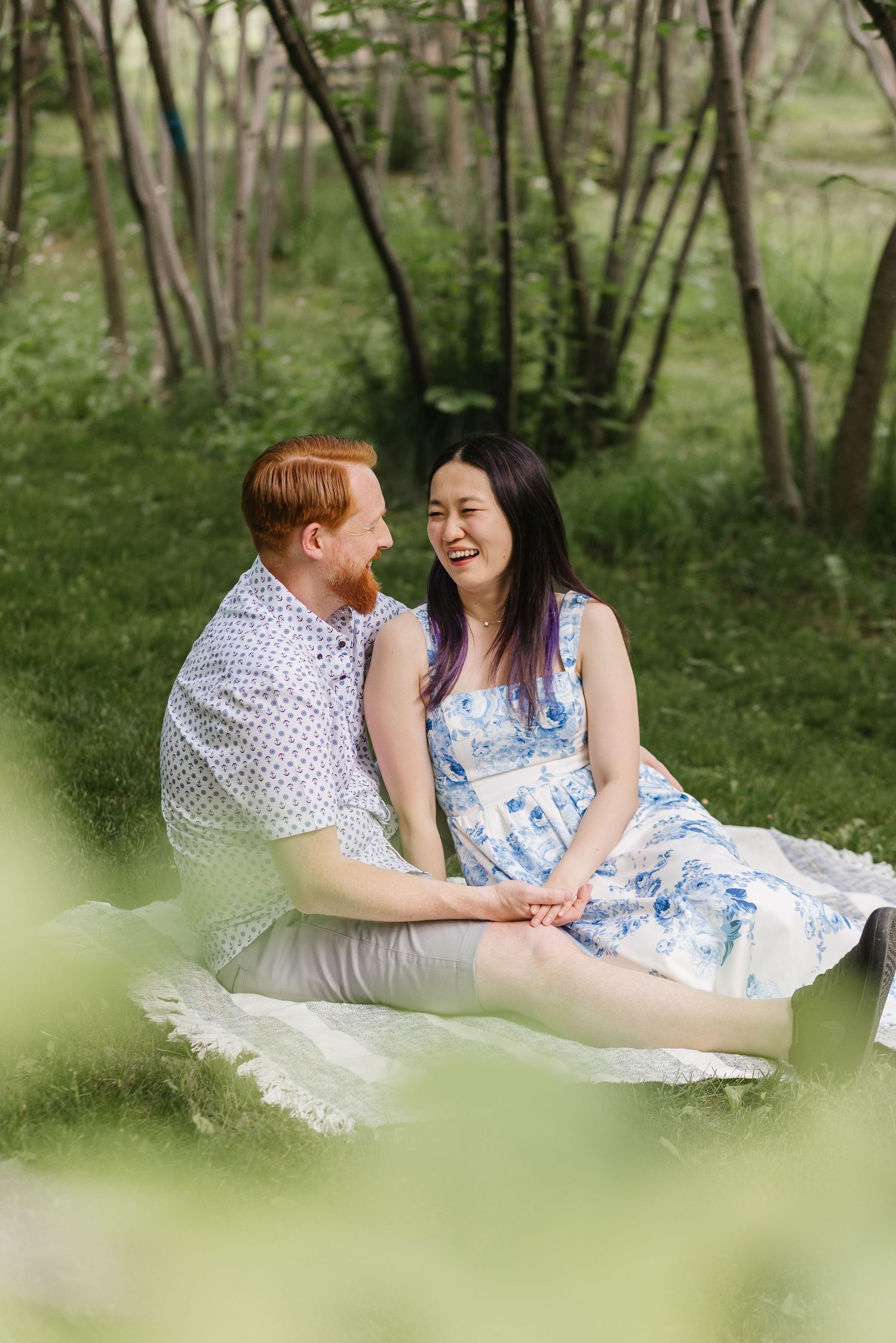 A couple sits together on a blanket in a wooded area, sharing a joyful moment among the trees and greenery.