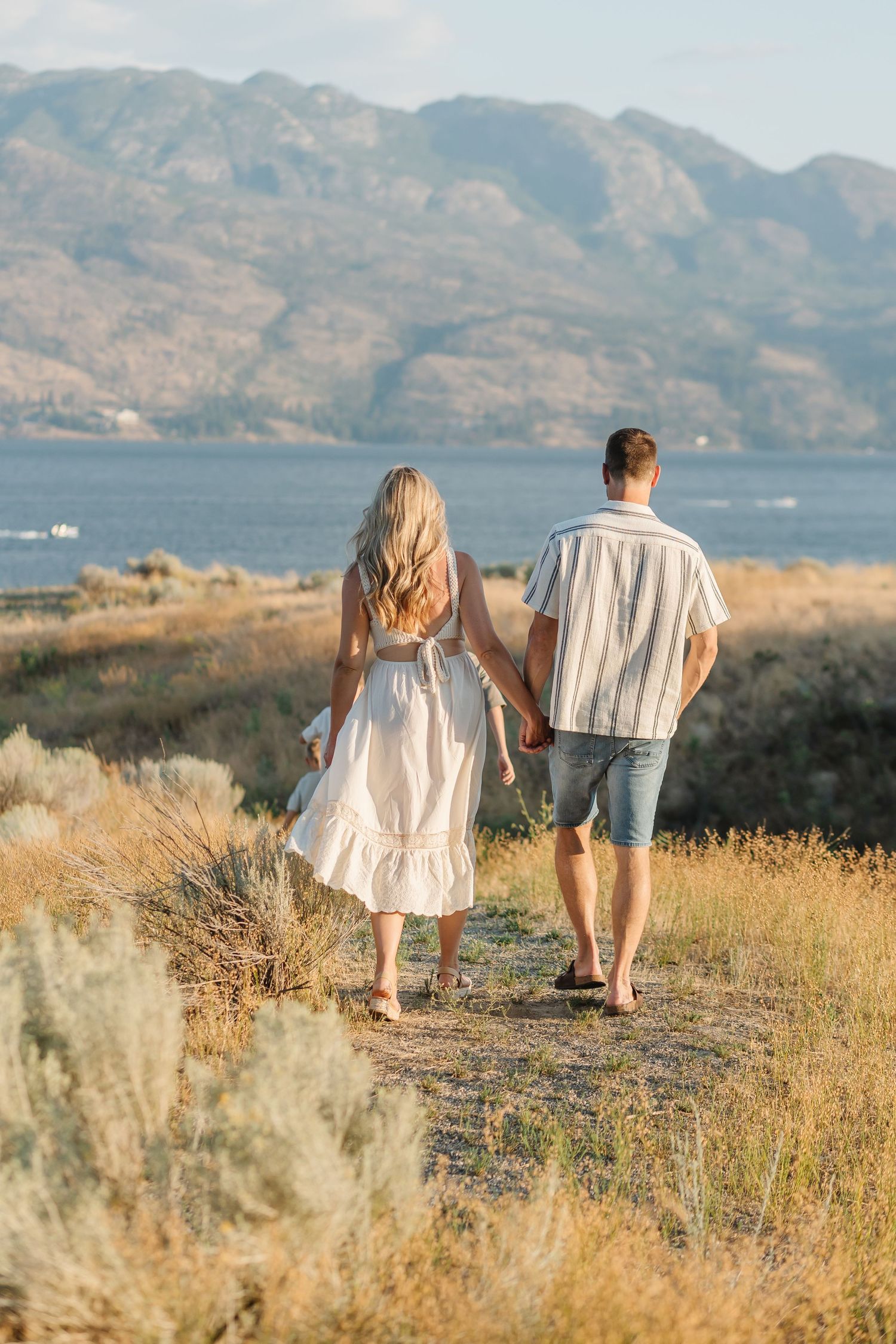A couple walks together on a scenic mountain trail through golden grass with a lake and mountains in the background.