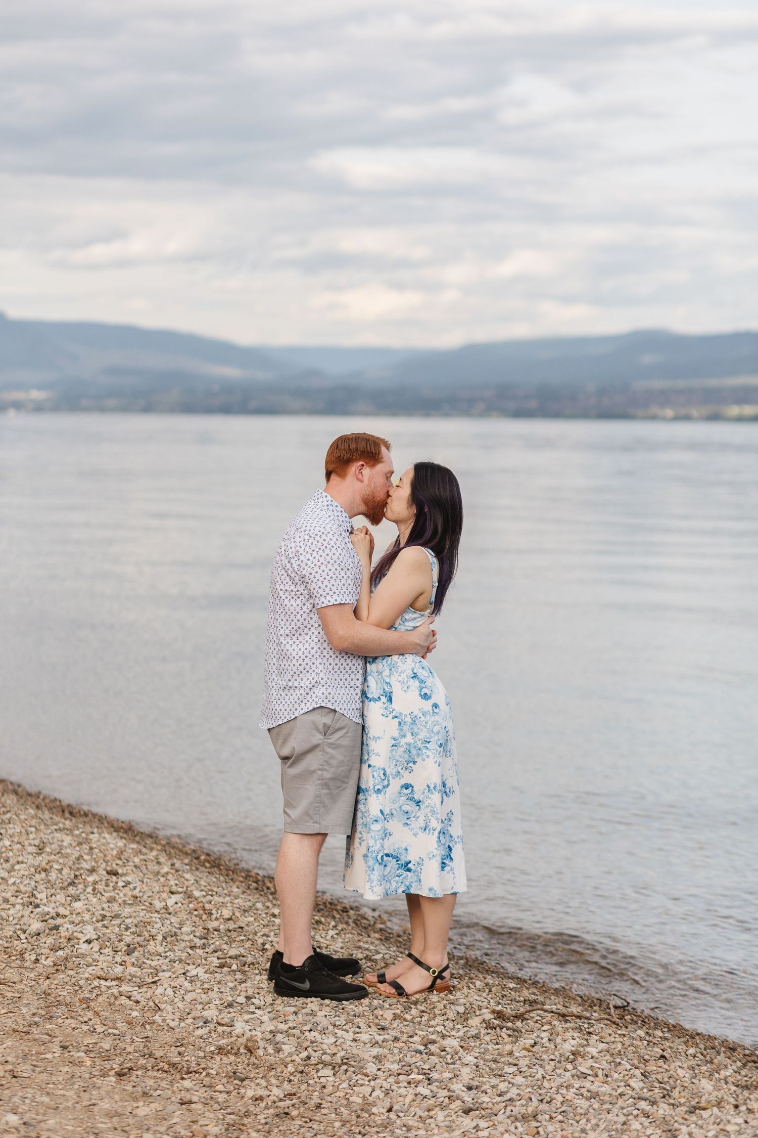 A couple shares a romantic moment on a pebble beach with mountains and a lake in the background.