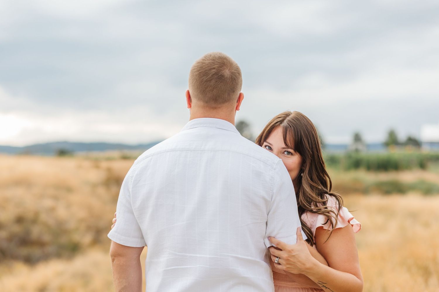 A couple in white outfits embraces while standing in a golden wheat field at sunset.