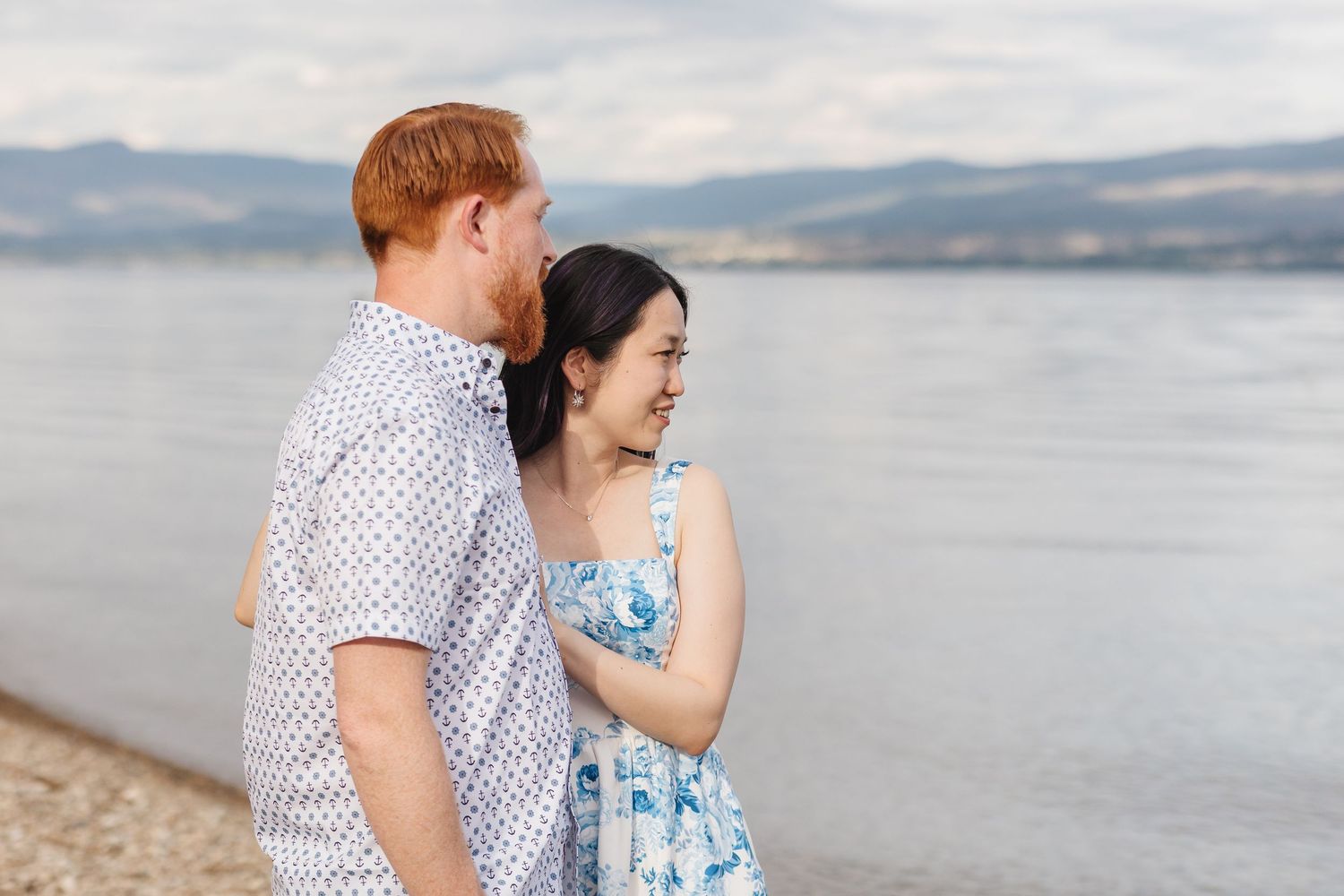 A couple embraces on a rocky beach with mountains and water in the background on a cloudy day.