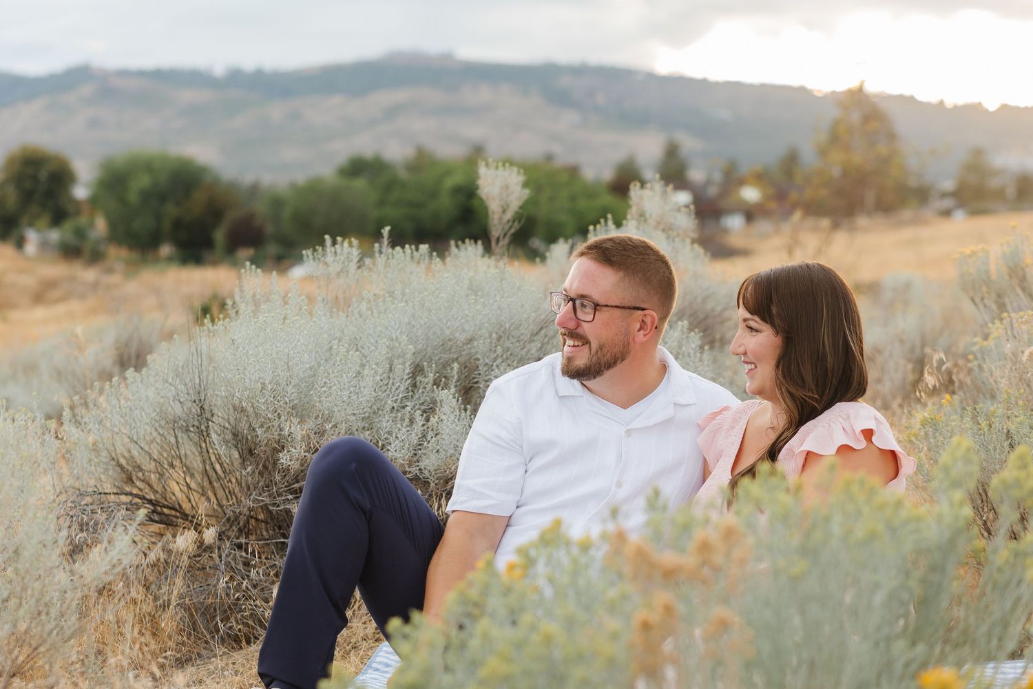 A couple sits together in a field of tall grass and sagebrush with rolling hills in the background during golden hour.