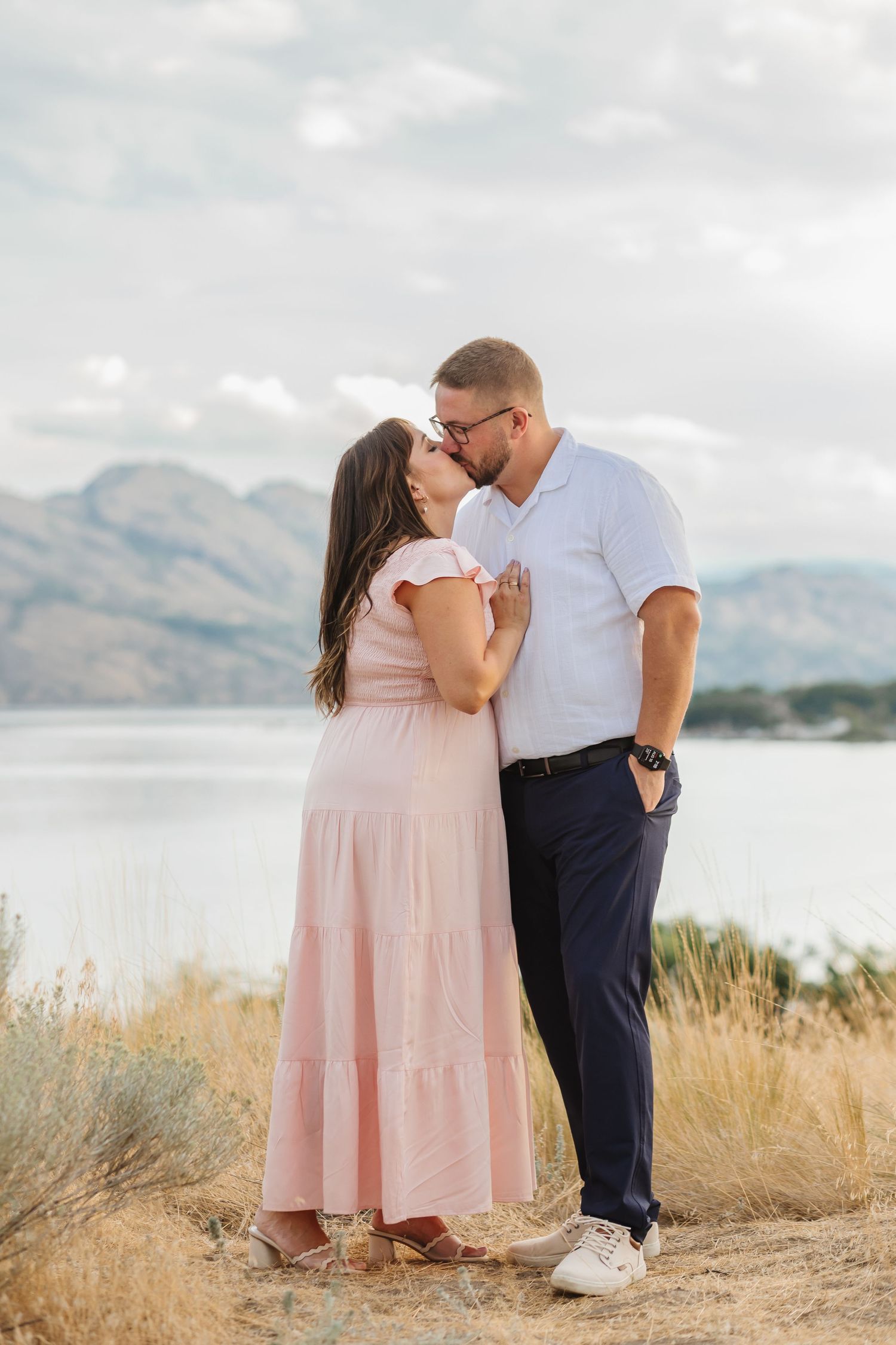 A couple shares an intimate moment against a scenic mountain lake backdrop with golden grass in the foreground.