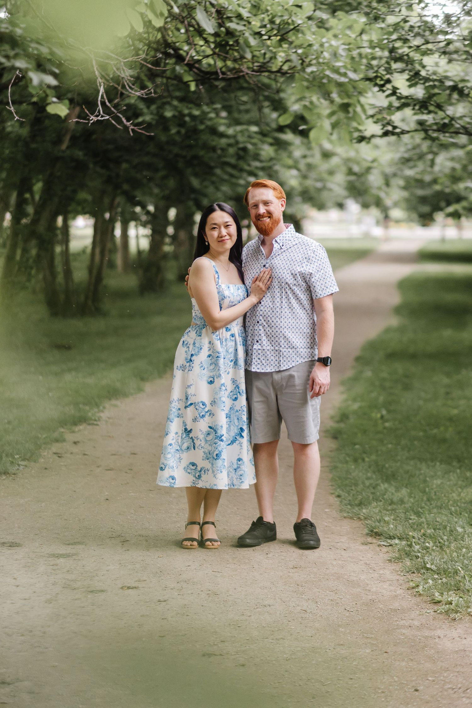A couple walking together on a tree-lined dirt path, with the woman wearing a blue floral dress and man in plaid shirt.