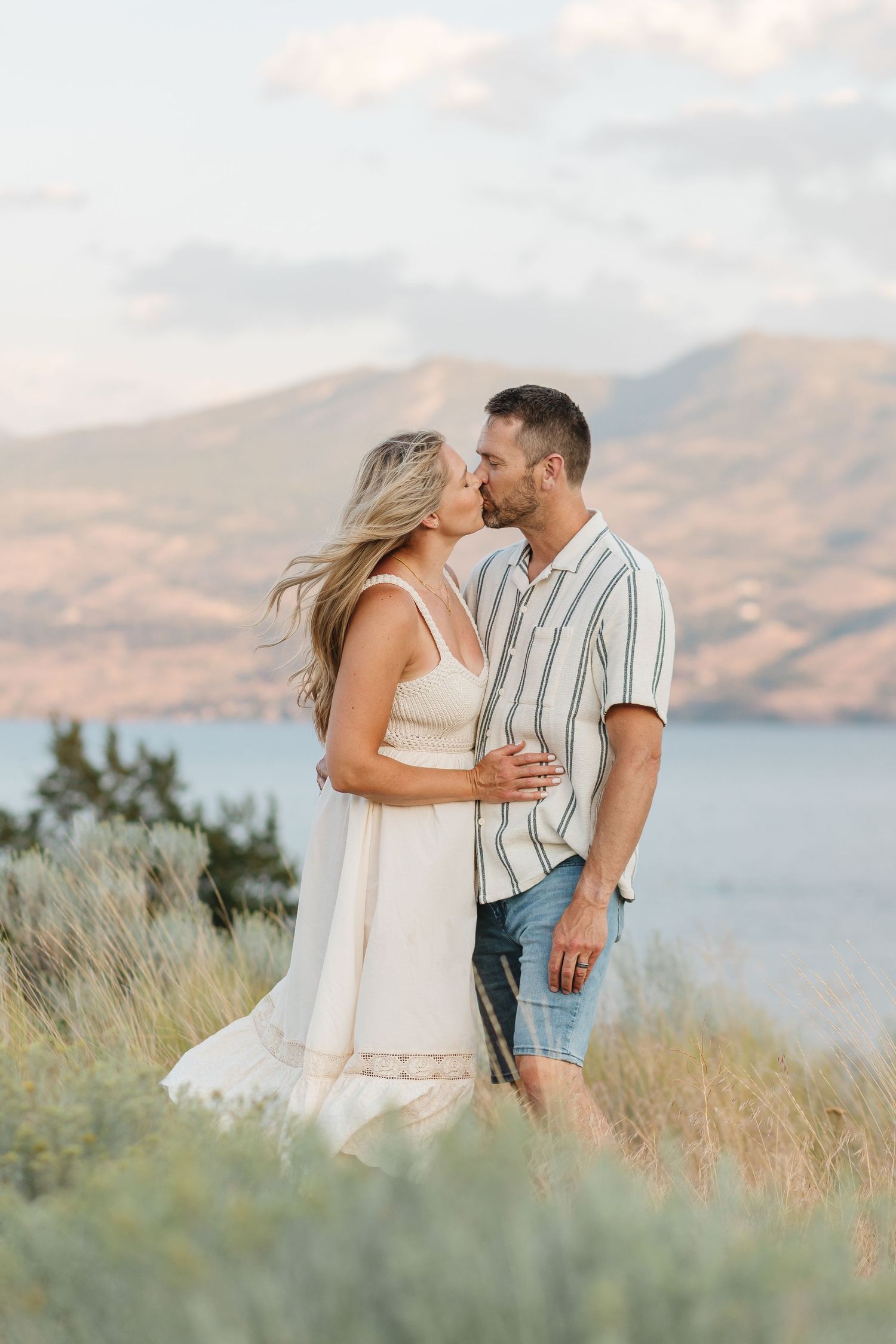 A romantic couple embraces on a hilltop overlooking a lake and mountains during golden hour.