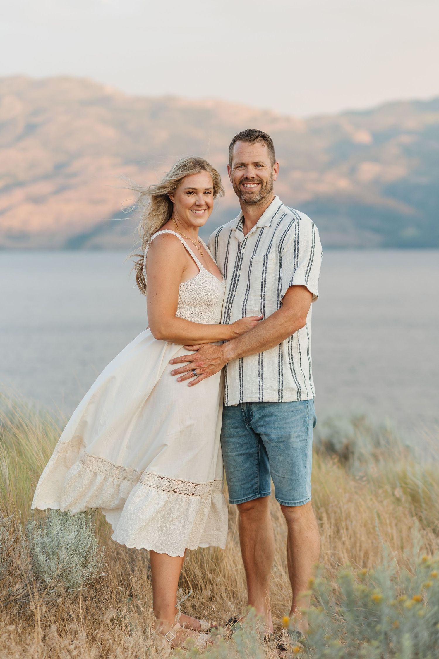 Couple embracing and laughing together in a grassy field with mountains and lake views behind them.