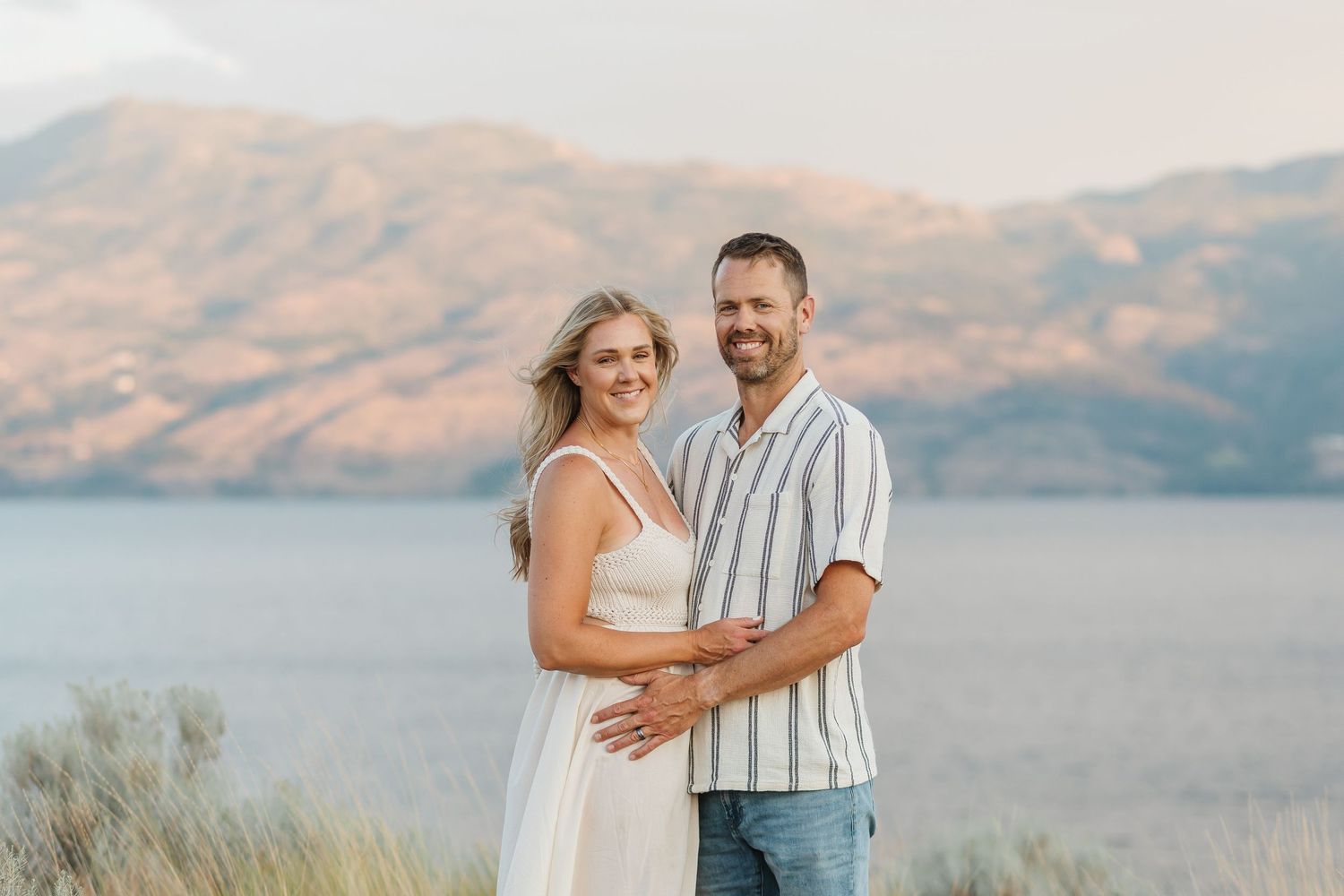 A couple embraces while standing on a hilltop overlooking a scenic mountain lake landscape at sunset.