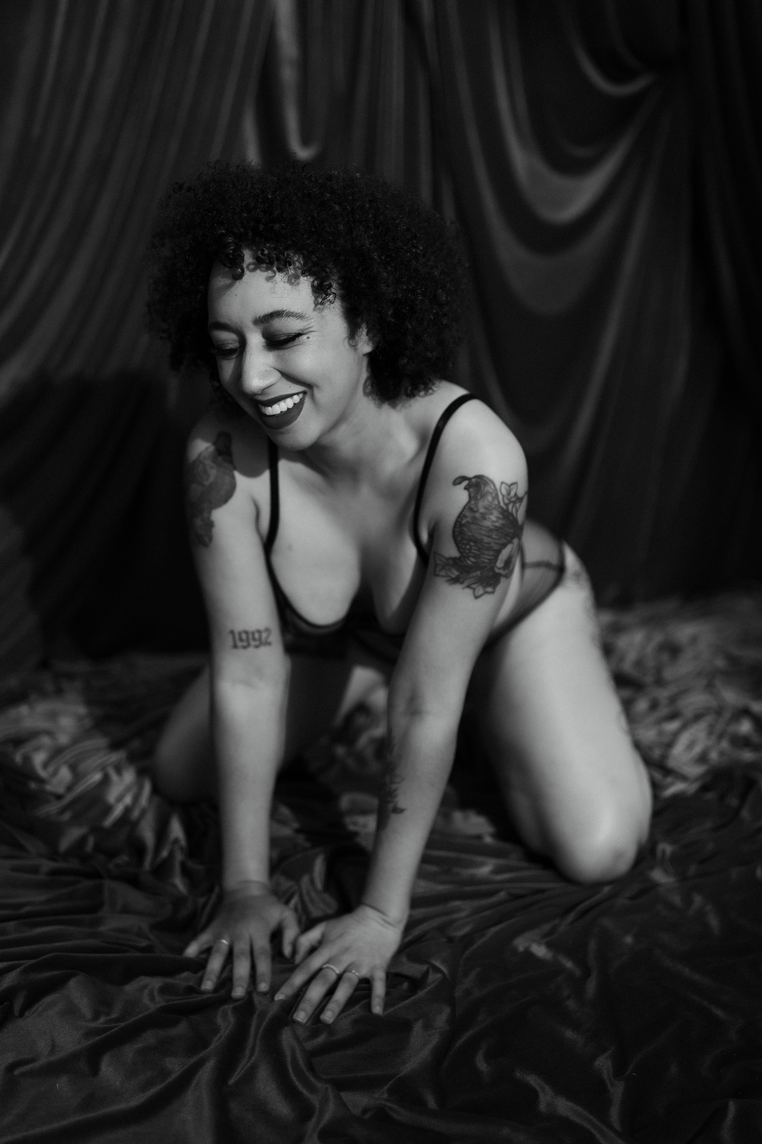 adult person kneeling on fabric backdrop, smiling while posing in black lingerie during a boudoir photo session