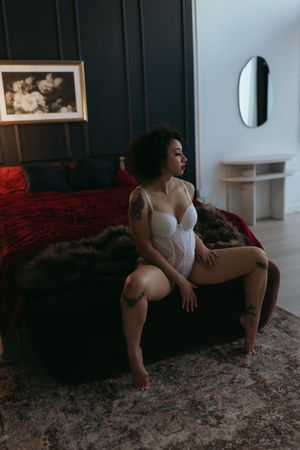 adult person sitting on a bench with fur, wearing light colored lingerie, posed with legs wide and looking away
