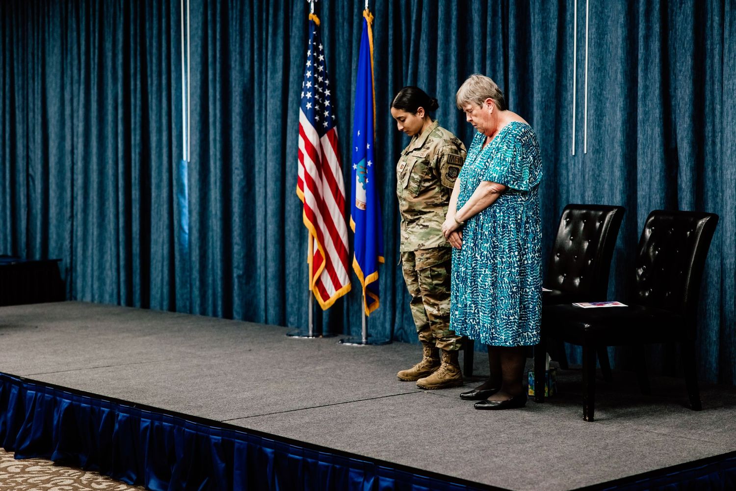 Military ceremony with uniformed service member and attendee against blue curtain backdrop.