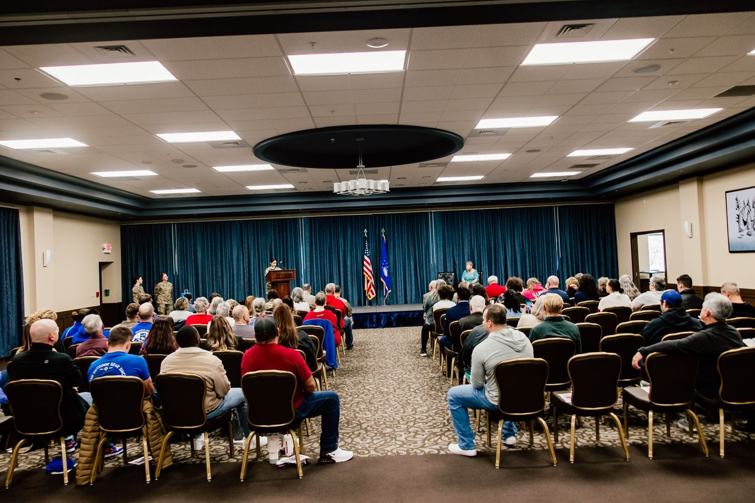 Conference room setup with rows of chairs and blue curtained stage with American flag.