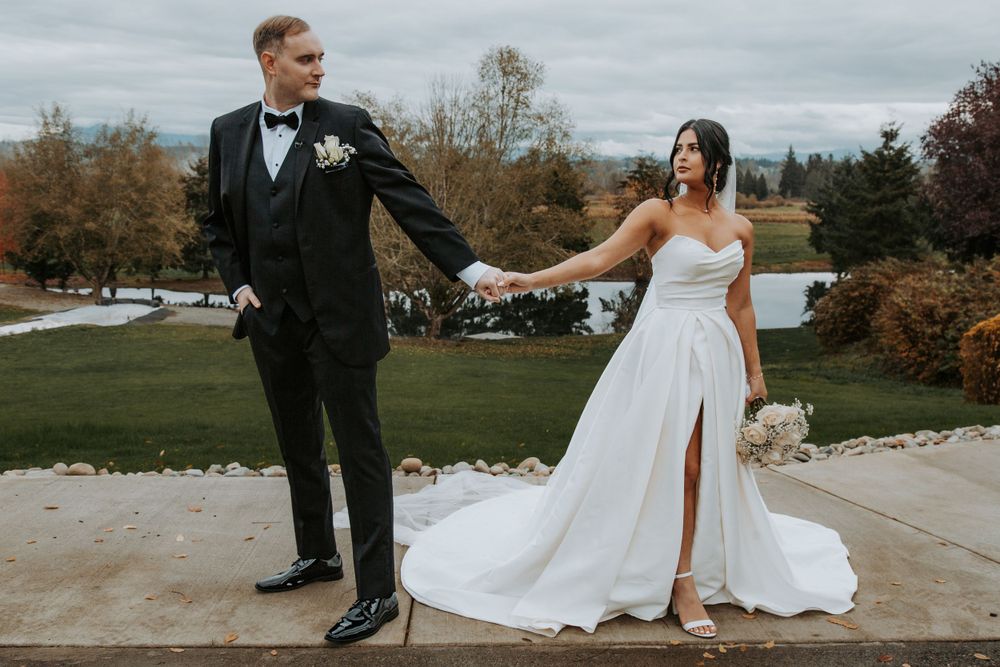Bride in a white split-front gown and groom in a black suit hold hands on a stone path at an autumn wedding venue.