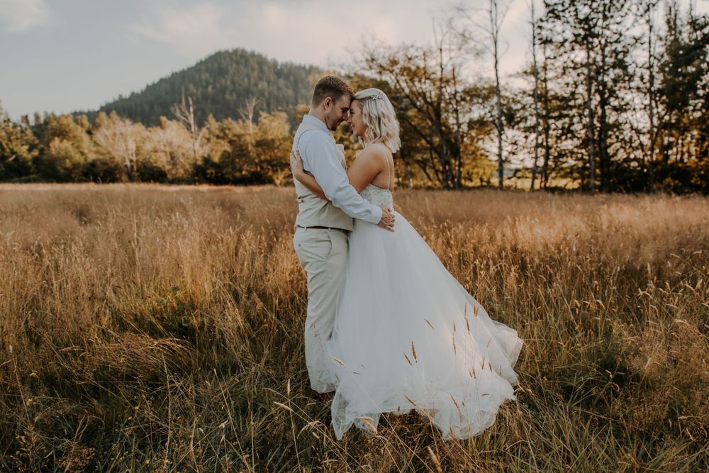 Bride and groom embrace foreheads together in a golden meadow with a forested mountain rising in the background.