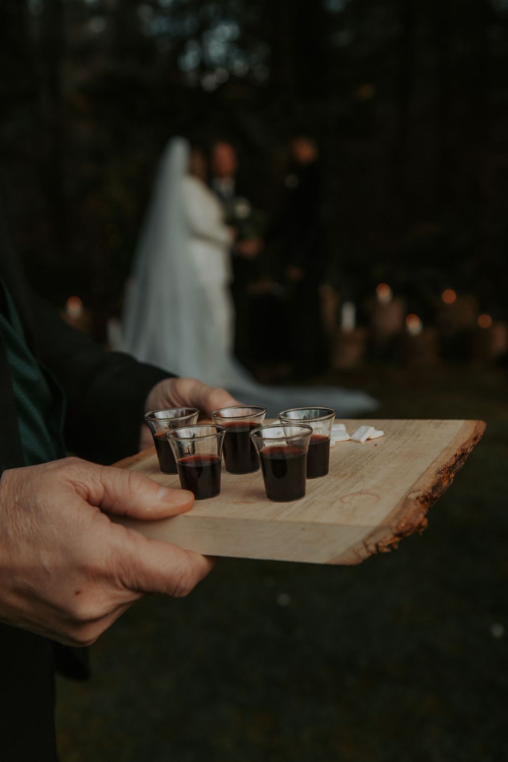 Hand holds a wooden tray of small cups of wine for a wedding communion with the veiled bride in the background.