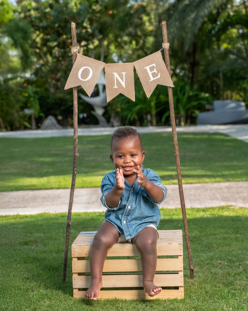 Adorable baby boy sitting on wooden crate under ONE banner sign celebrating his first birthday outdoors.
