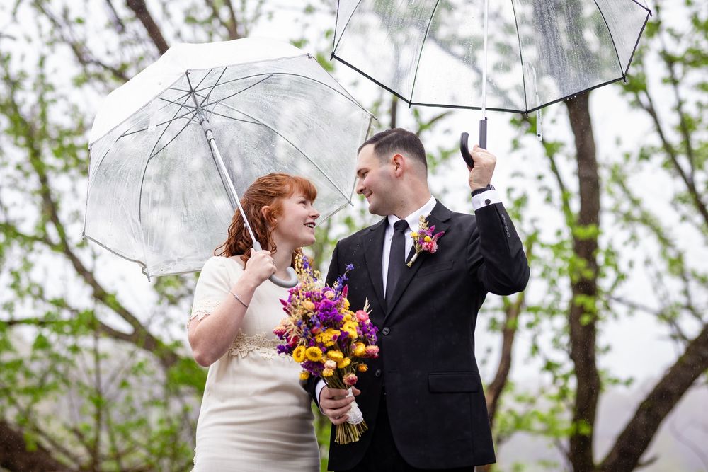 Bride and groom holding clear umbrellas in the rain, bride carrying colorful bouquet, sharing a romantic glance outdoors.