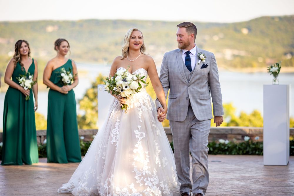 Bride and groom walk hand-in-hand at outdoor wedding ceremony with bridesmaids in emerald green dresses.