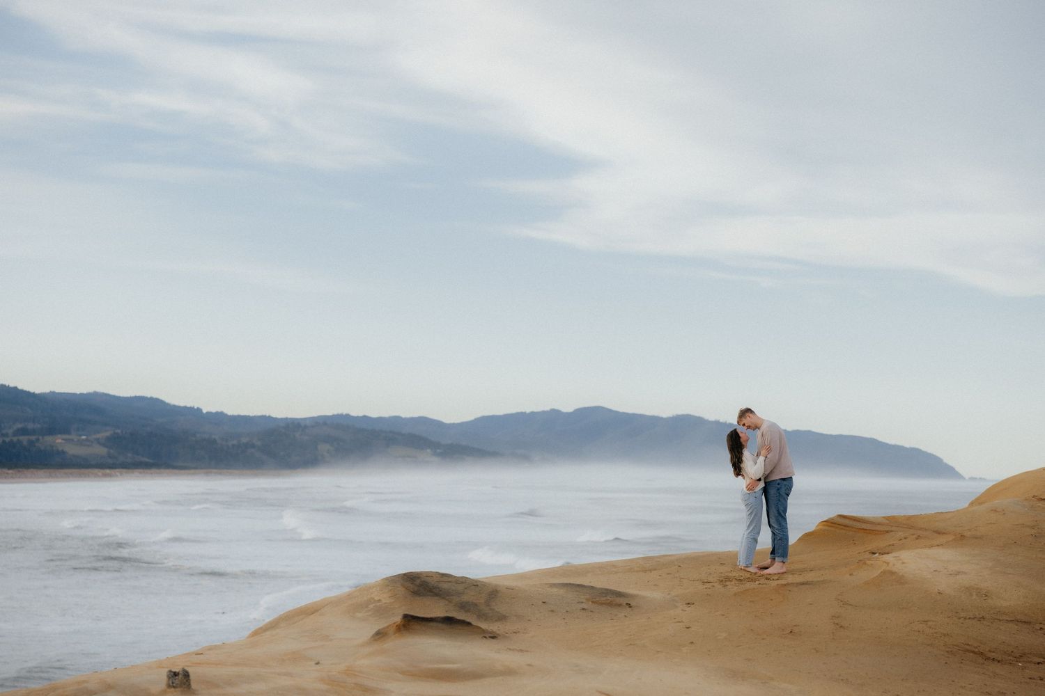 Couple stands on sandy cliff overlooking misty coastal mountain landscape during sunset at Oregon Coast.