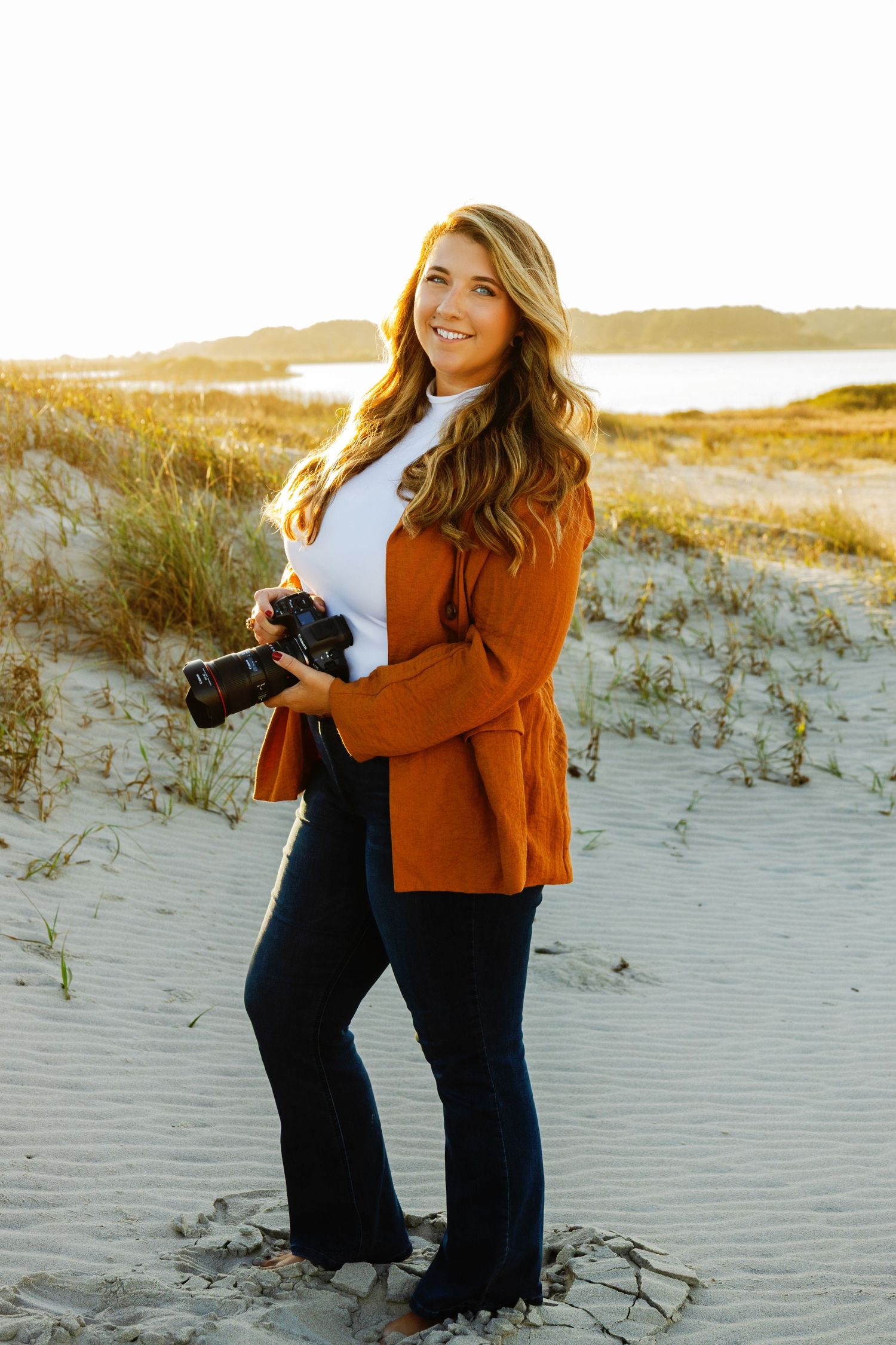 Smiling female photographer holding a camera on a sandy beach at golden hour in an orange blazer.