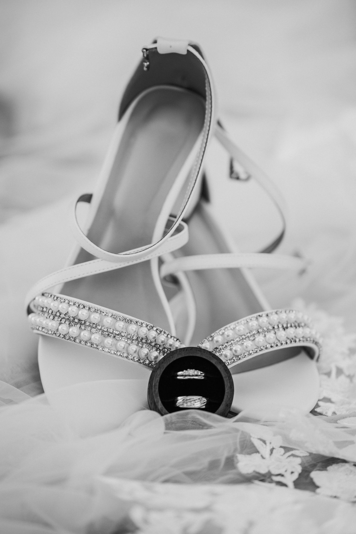 Black and white photo of elegant bridal ballet flats with rhinestone details and wedding rings displayed on tulle fabric.
