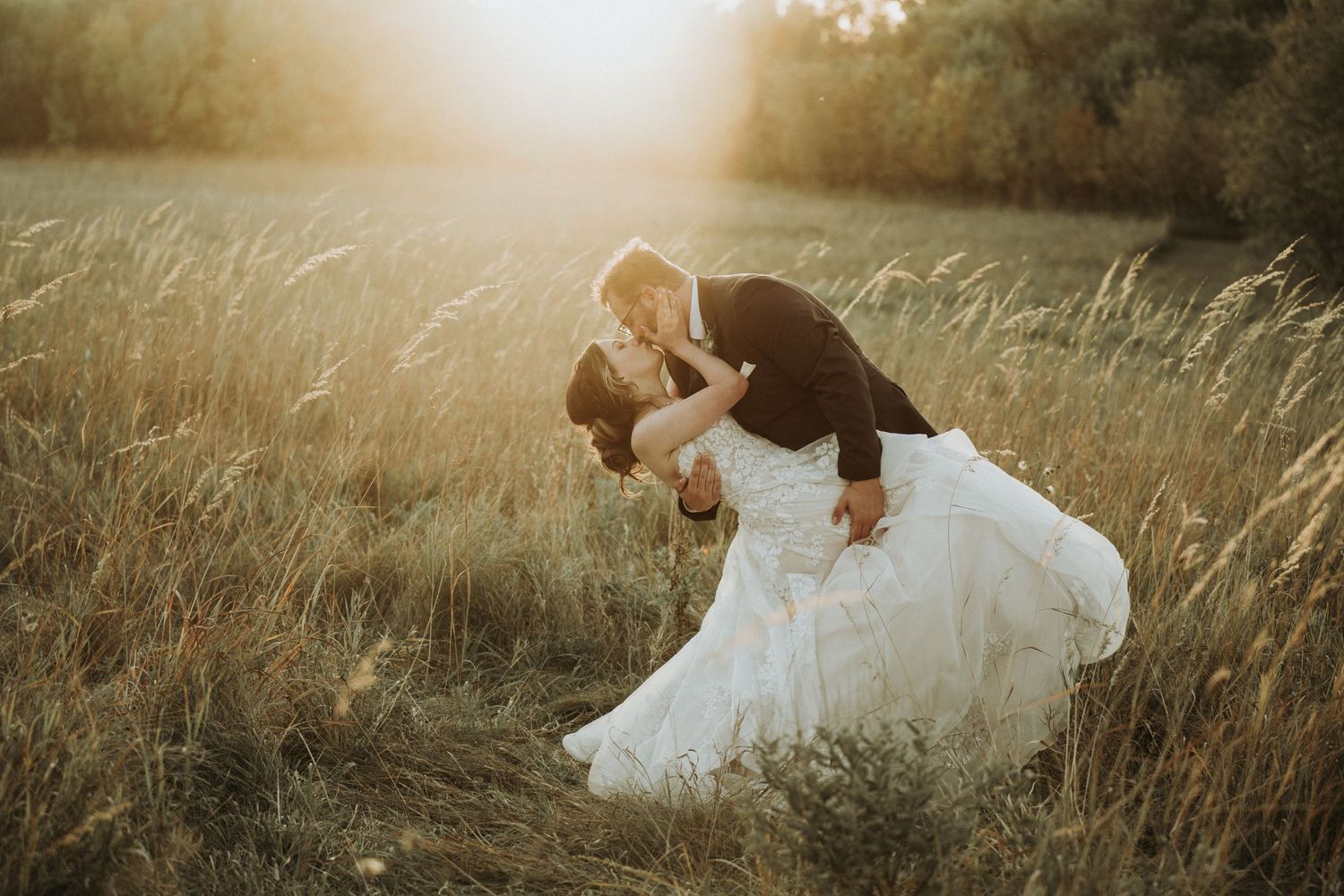 A romantic couple in formal wedding attire share an intimate moment in a sunlit meadow at golden hour.