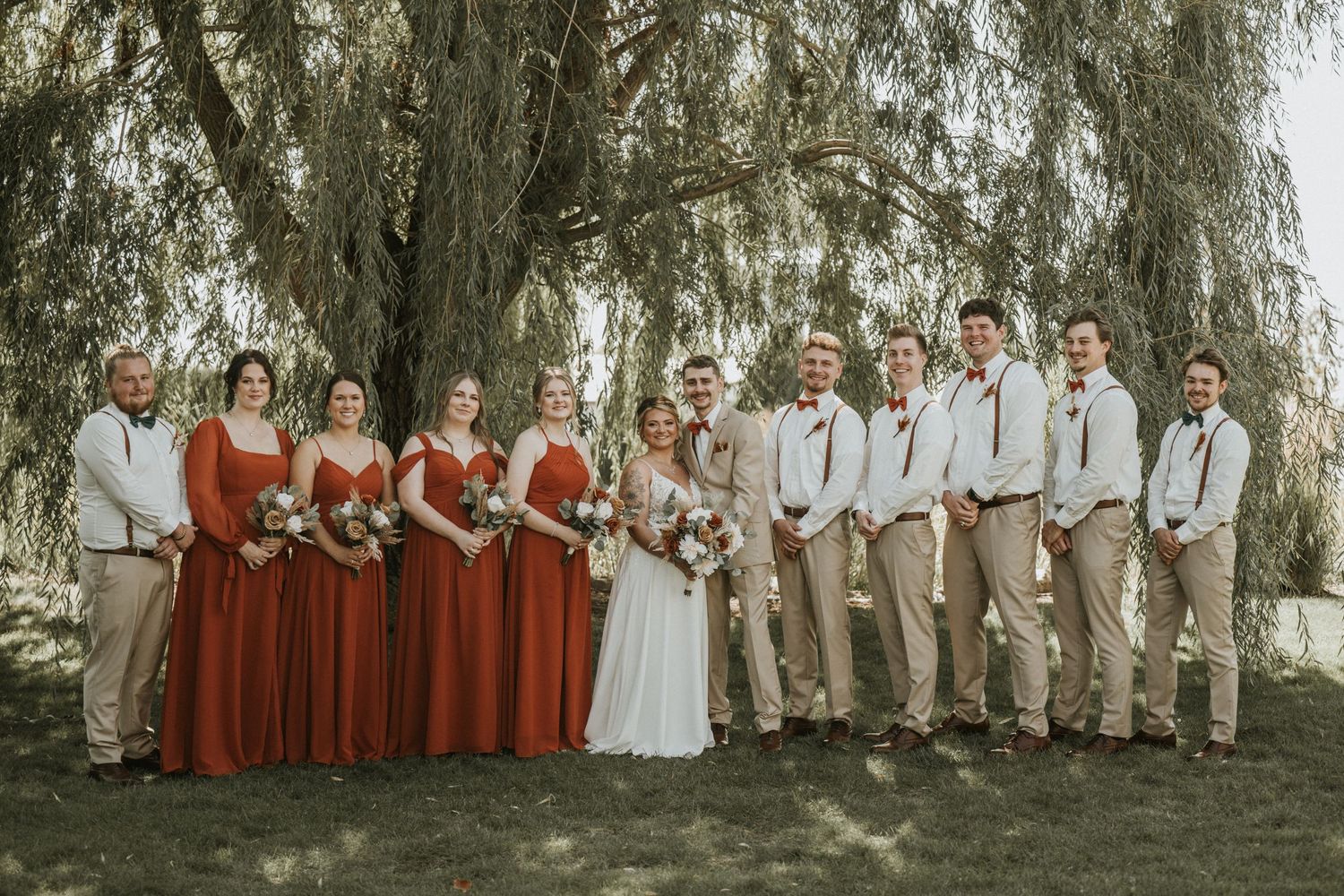 Wedding party in burgundy dresses and khaki suits poses under drooping willow tree branches in natural outdoor setting.