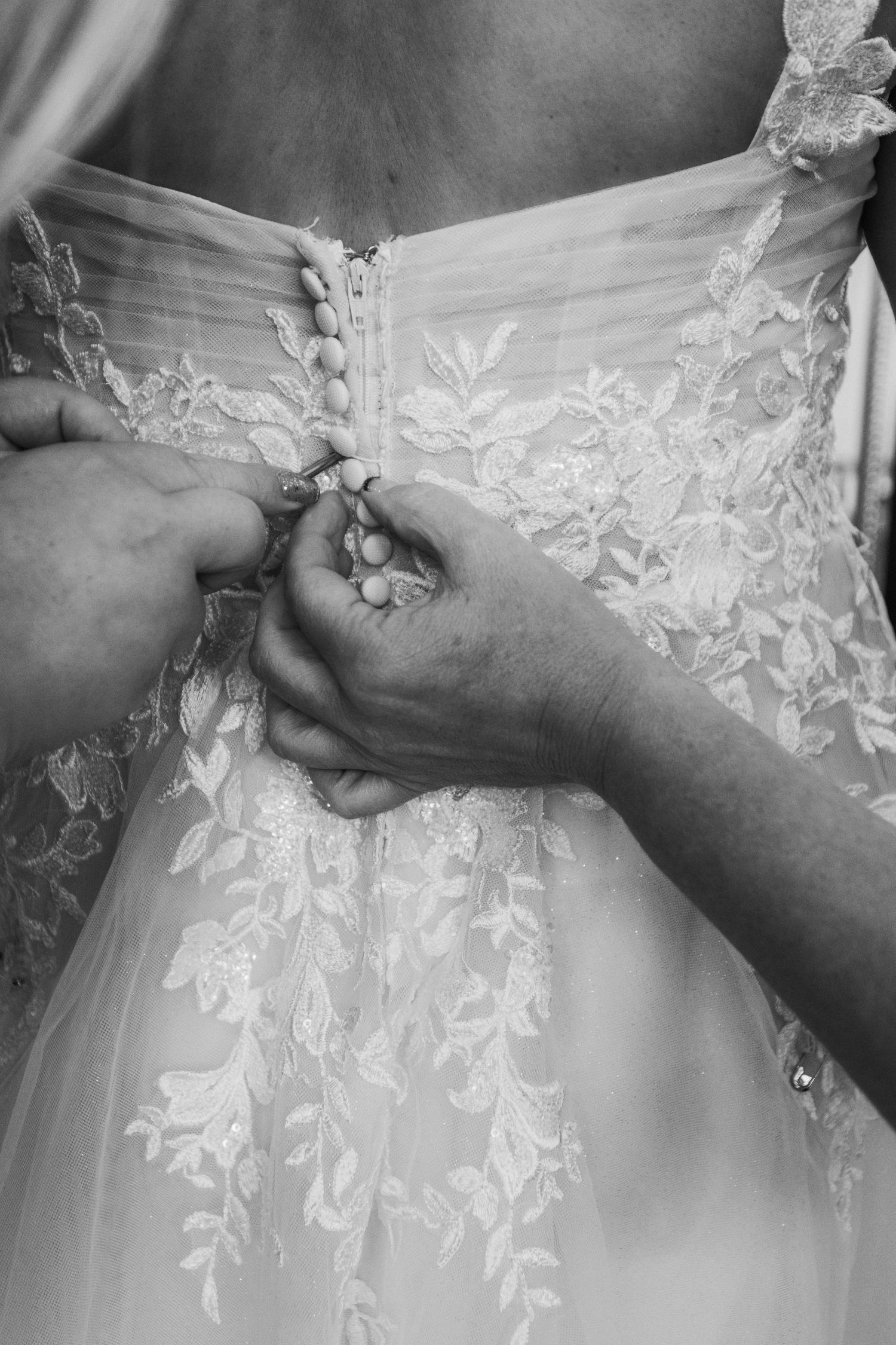 Black and white close-up of hands fastening delicate buttons on an ornate lace wedding dress with floral embroidery.