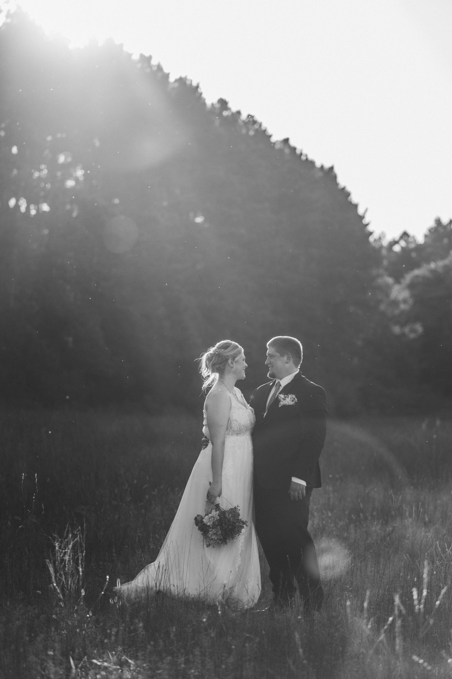Black and white wedding photograph of a couple standing in a sunlit meadow with trees in the background.