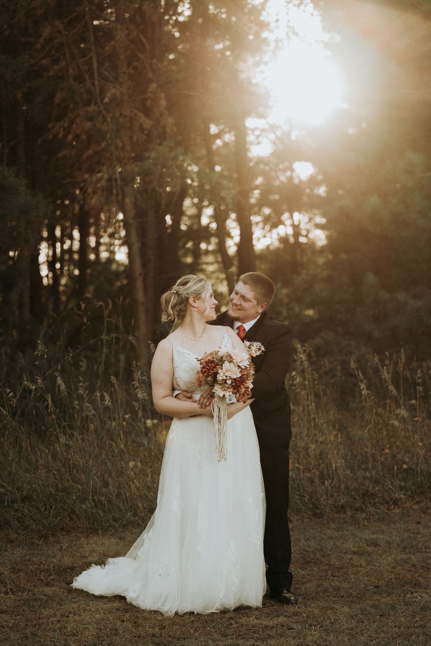 A romantic wedding couple shares tender moments at sunset in a wooded field with golden sunlight filtering through the trees.