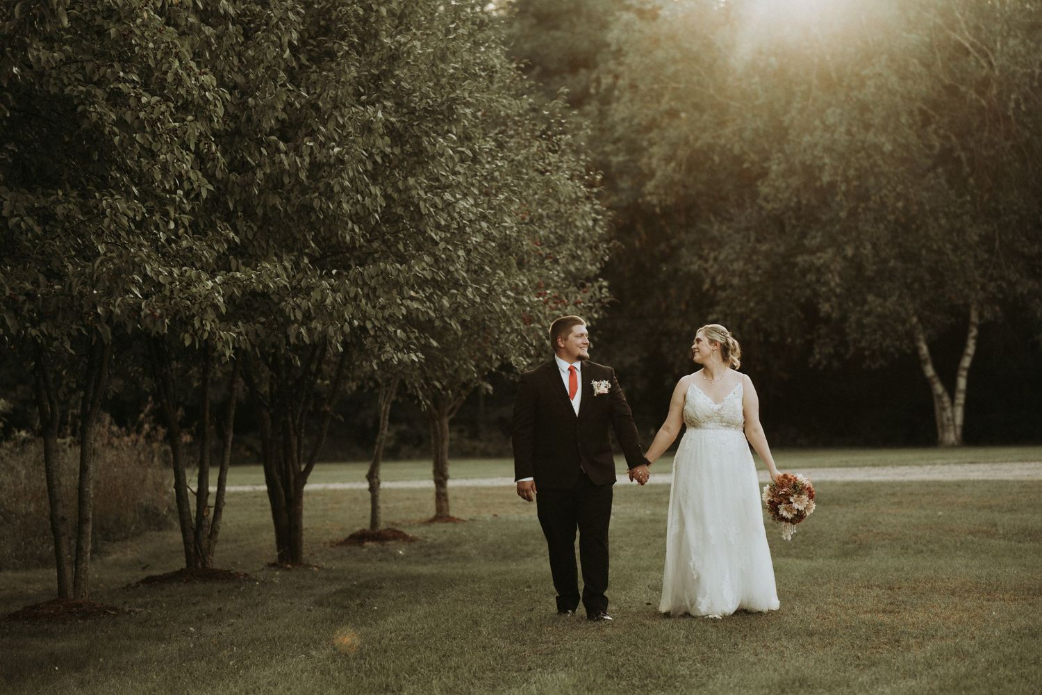 A couple walks hand in hand through a sunlit park on their wedding day as golden light filters through the trees.