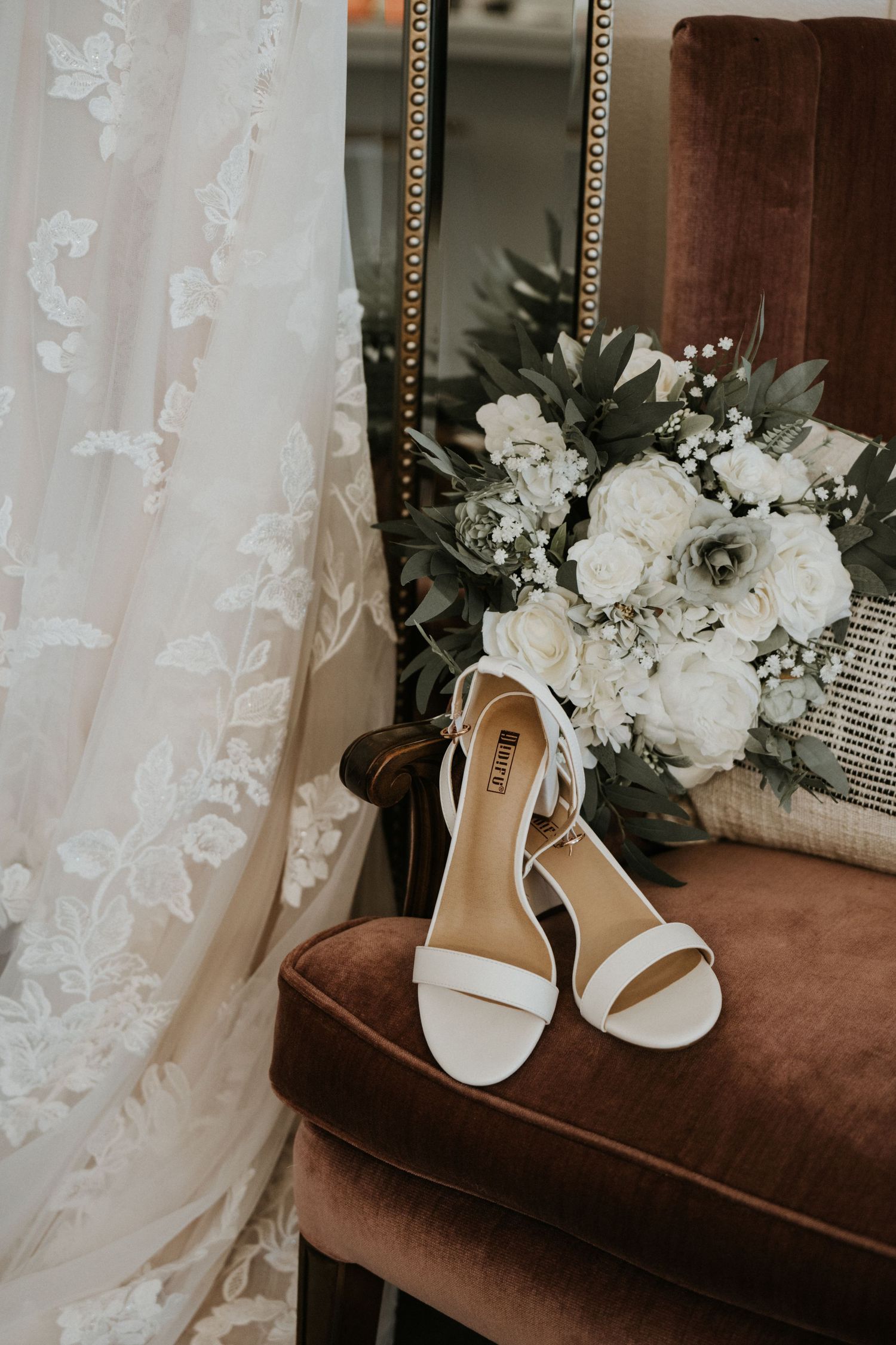 White bridal sandals and floral bouquet displayed on brown velvet chair near lace wedding dress.