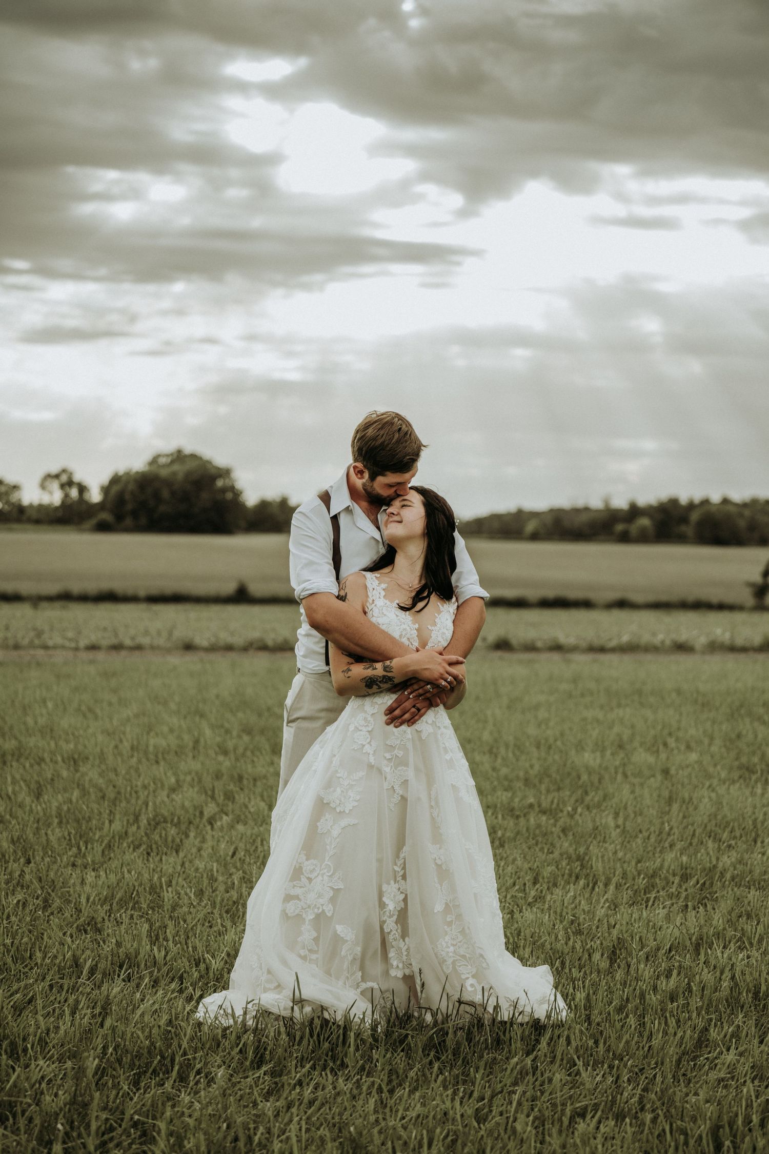 A couple embraces in a rustic outdoor field setting with dramatic cloudy sky during their wedding day. Minnesota wedding photographer