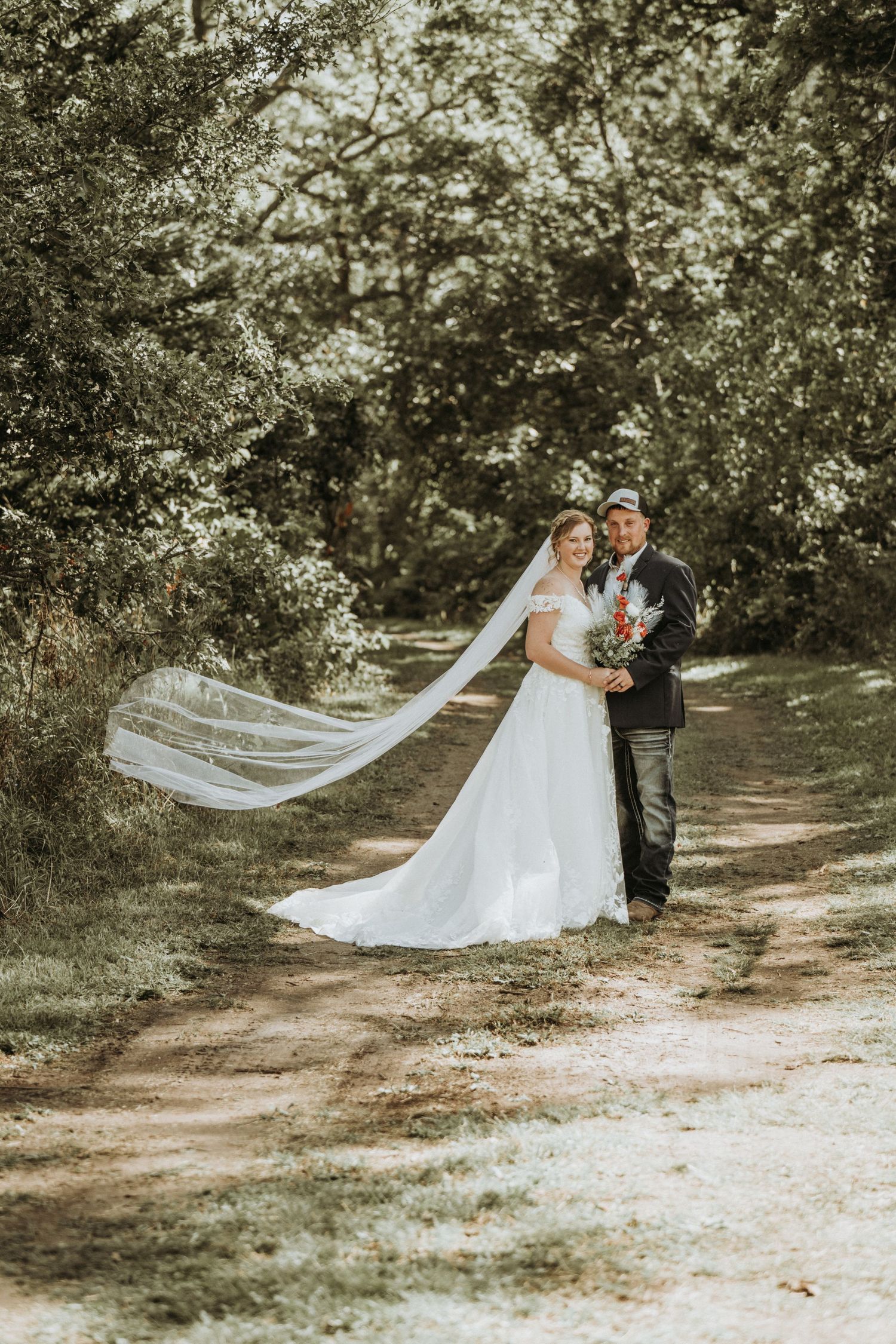 A romantic outdoor wedding photo showing newlyweds on a wooded path with a flowing long veil catching the breeze.