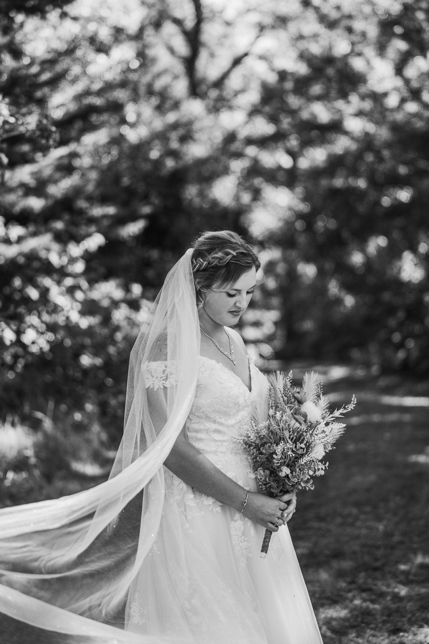 A bride in a flowing white gown and veil holds a rustic bouquet while standing in a sunlit garden setting.