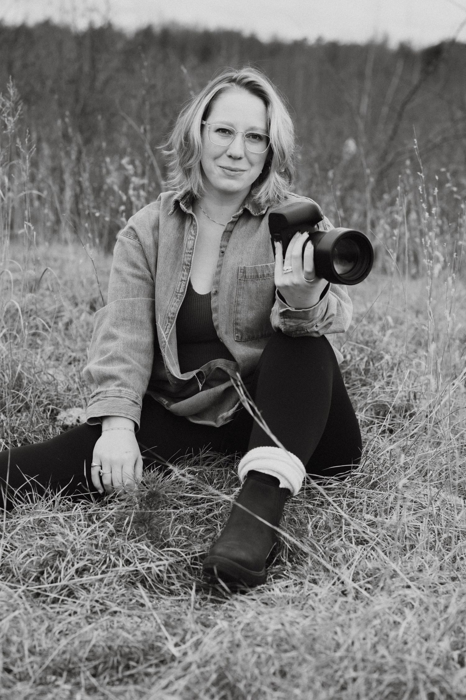 Black and white portrait of blonde female photographer sitting in field holding DSLR camera.