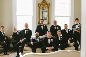 Group of groomsmen in black tuxedos lounging on elegant white furniture.