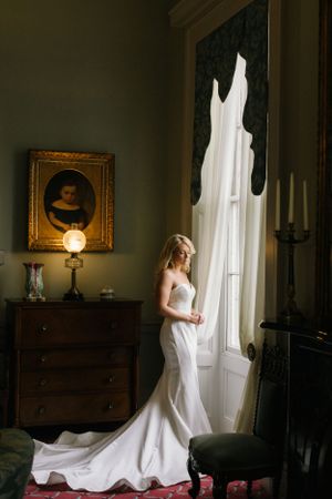Elegant white wedding dress displayed in ornate room with antique furniture and dramatic lighting.