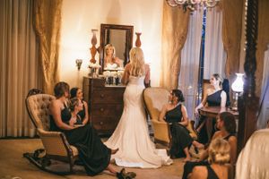 Wedding scene with bride in fitted gown surrounded by attendants in dark dresses.