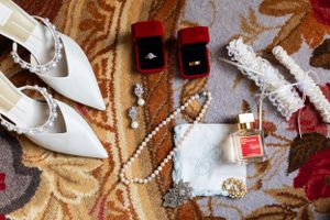 Wedding accessories including white heels, pearl necklace, rings and perfume arranged on patterned carpet.