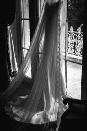 Artistic black and white photo of wedding dress train flowing by window.
