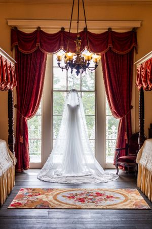 White wedding gown hanging in elegant room with red curtains and ornate carpet.