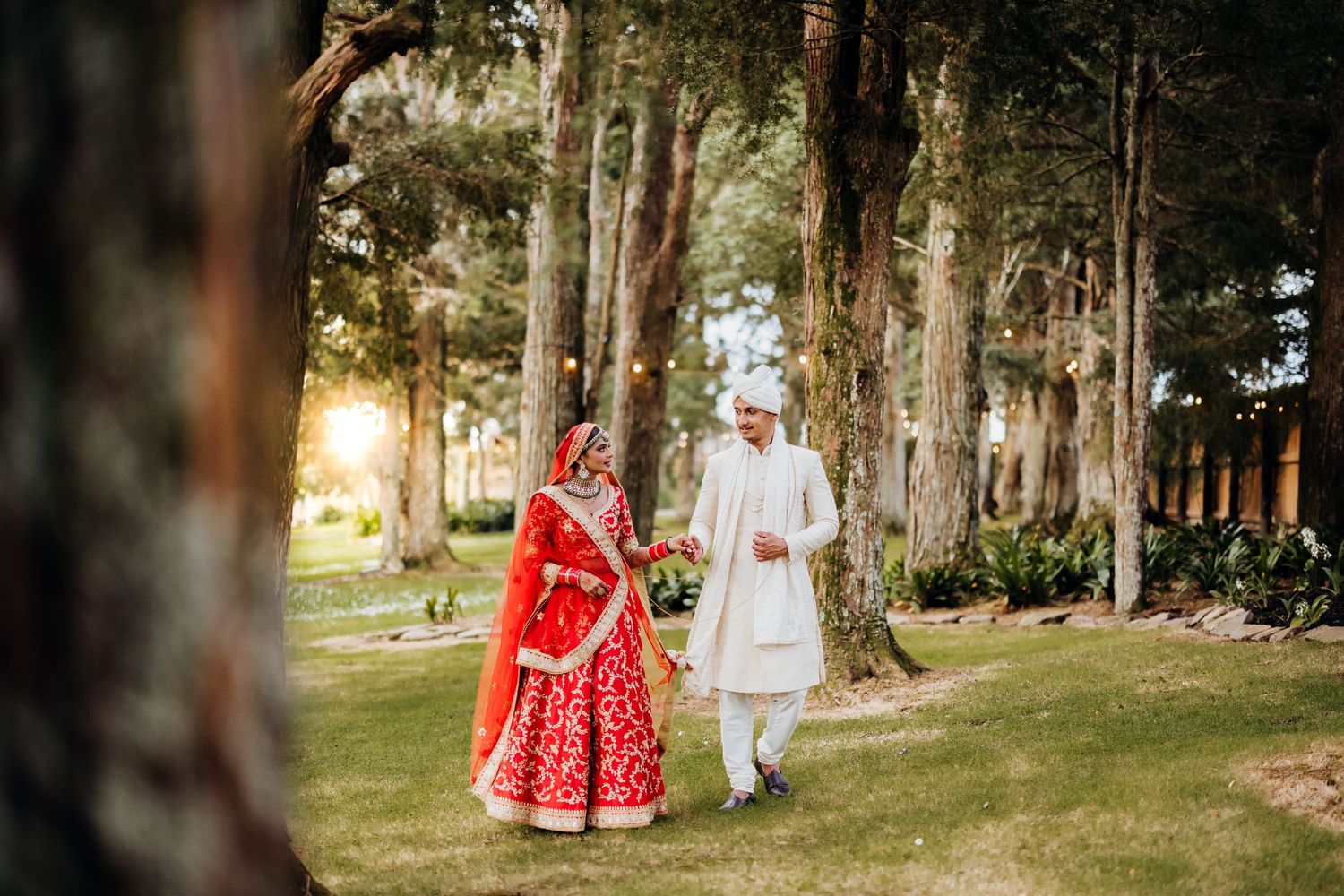 Indian bride in red lehenga and groom in white sherwani walking hand in hand through a sunlit forest.
