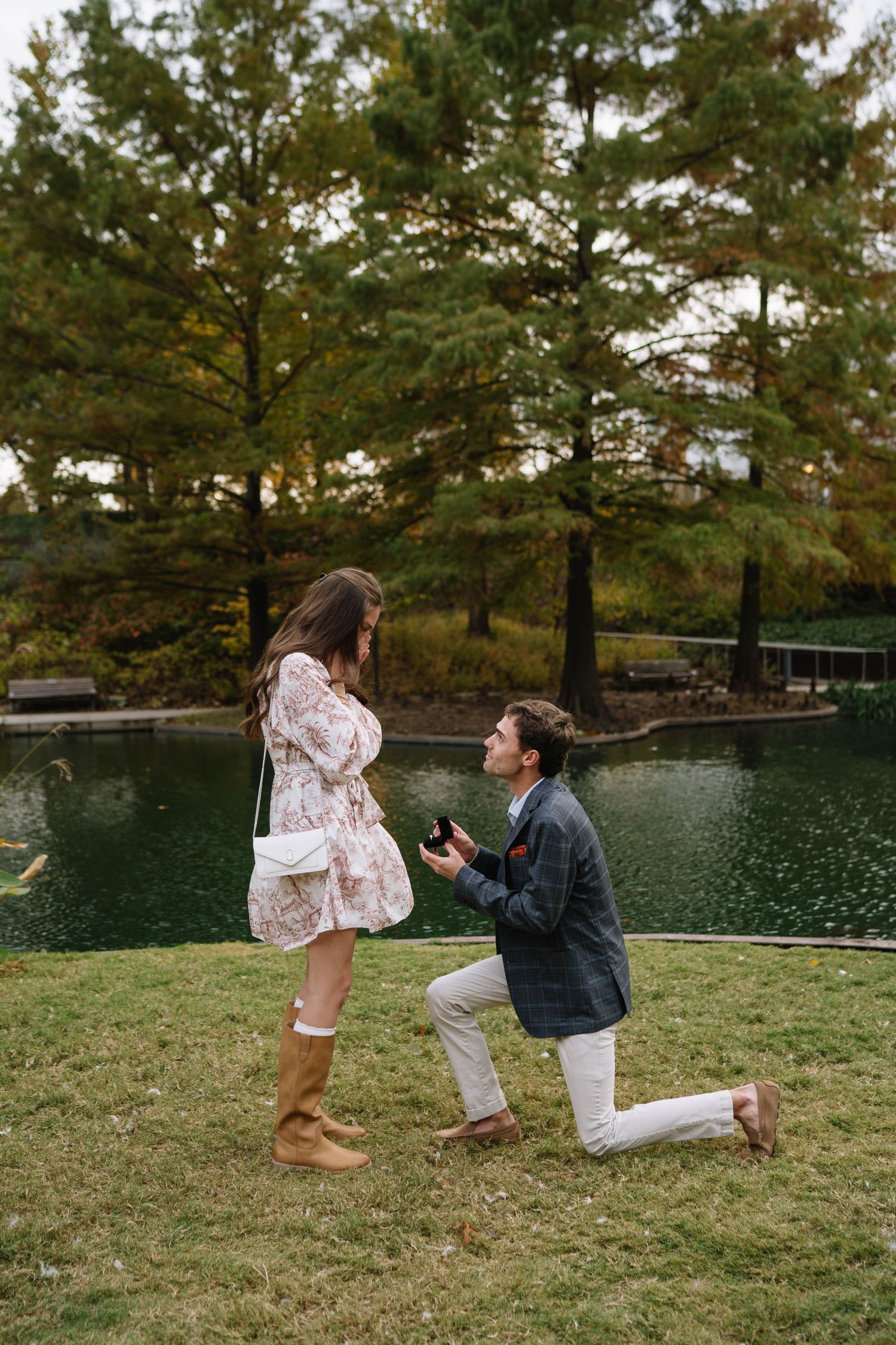 A couple shares a romantic proposal moment by a serene lake surrounded by pine trees at sunset.