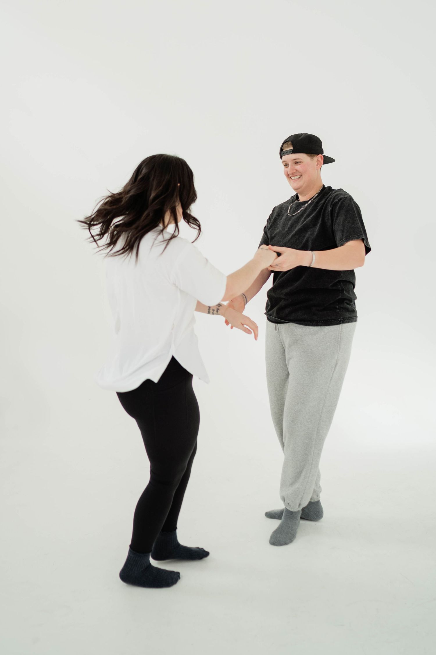 Two people dancing together in a studio, one wearing a white shirt and black leggings, the other in a black tee and gray sweatpants.