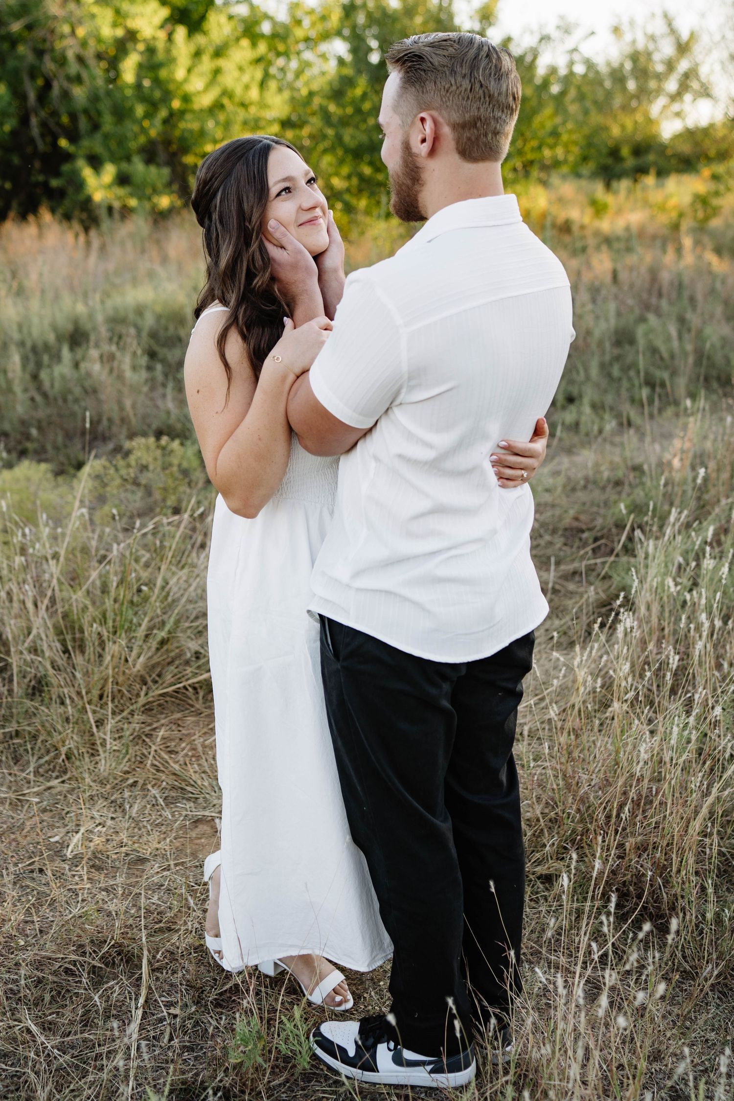 A couple in white and black outfits embrace lovingly in a golden sunlit field of tall grass.