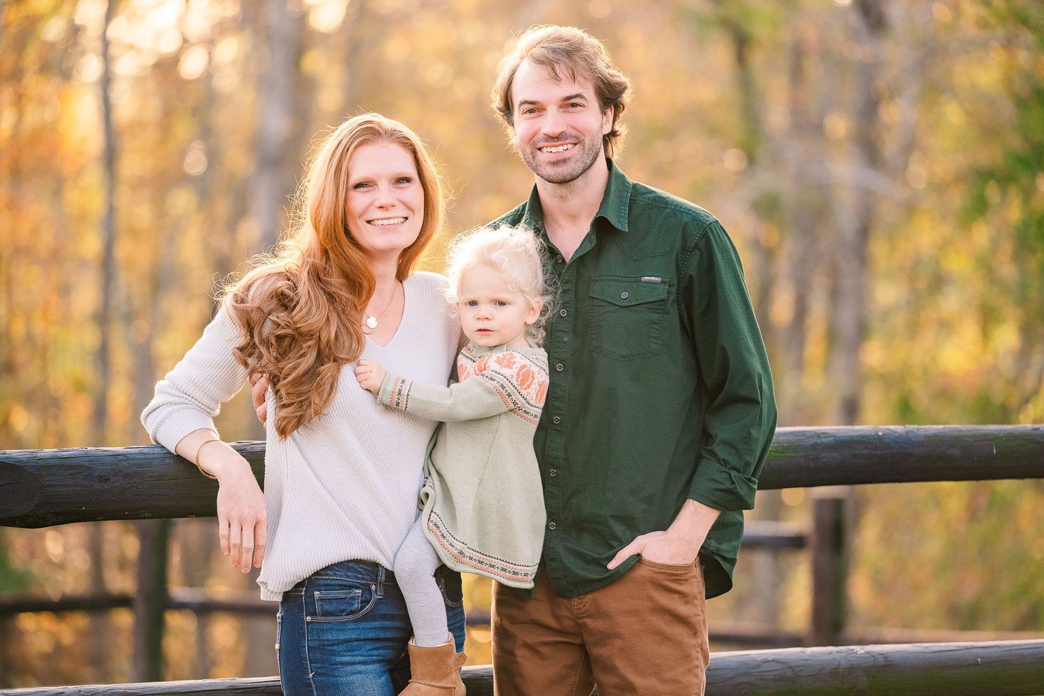A happy young family of three poses on a wooden bridge surrounded by colorful autumn foliage.
