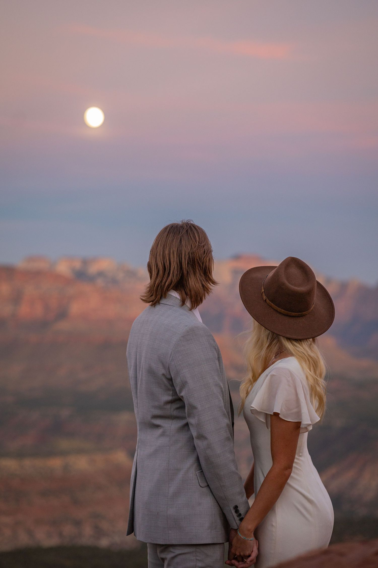A couple stands on a red rock cliff overlooking mountains at sunset with a full moon rising in the background.