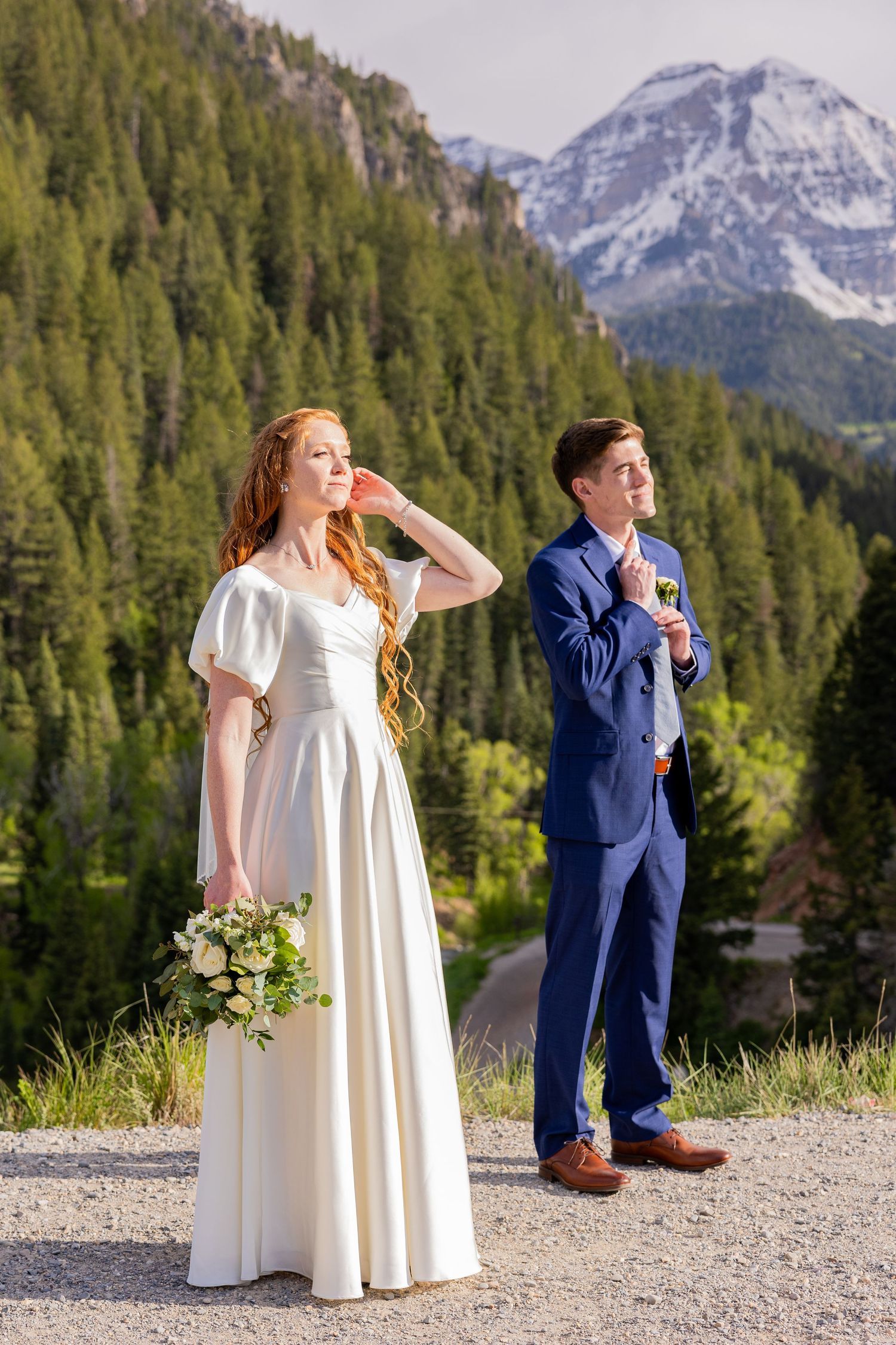Couple in wedding attire pose playfully against mountain landscape backdrop.